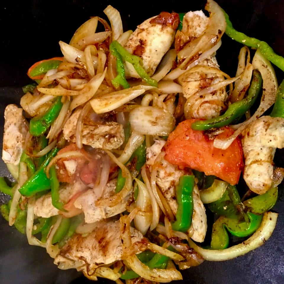 Sizzling chicken fajitas with grilled onions, green peppers, and red tomatoes on a dark surface.