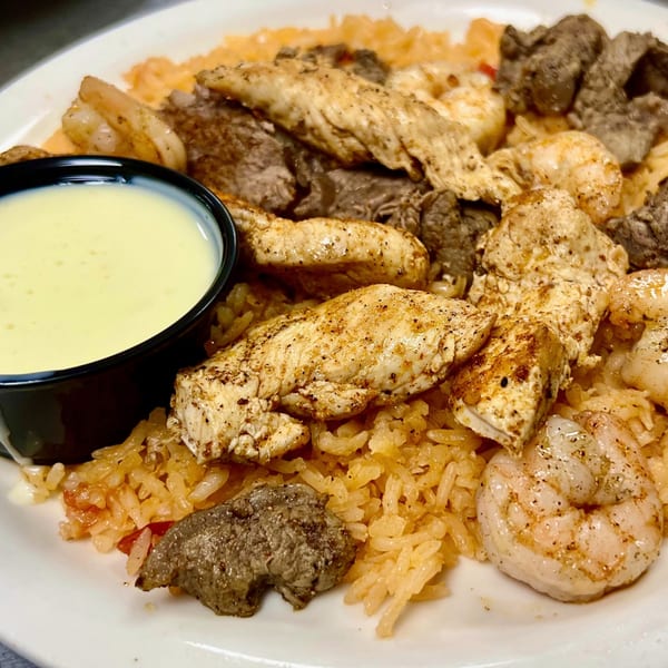 A plate of chicken, steak, and shrimp over rice, with a side of white sauce.