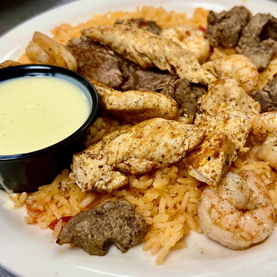 A plate of chicken, steak, and shrimp over rice, with a side of white sauce.