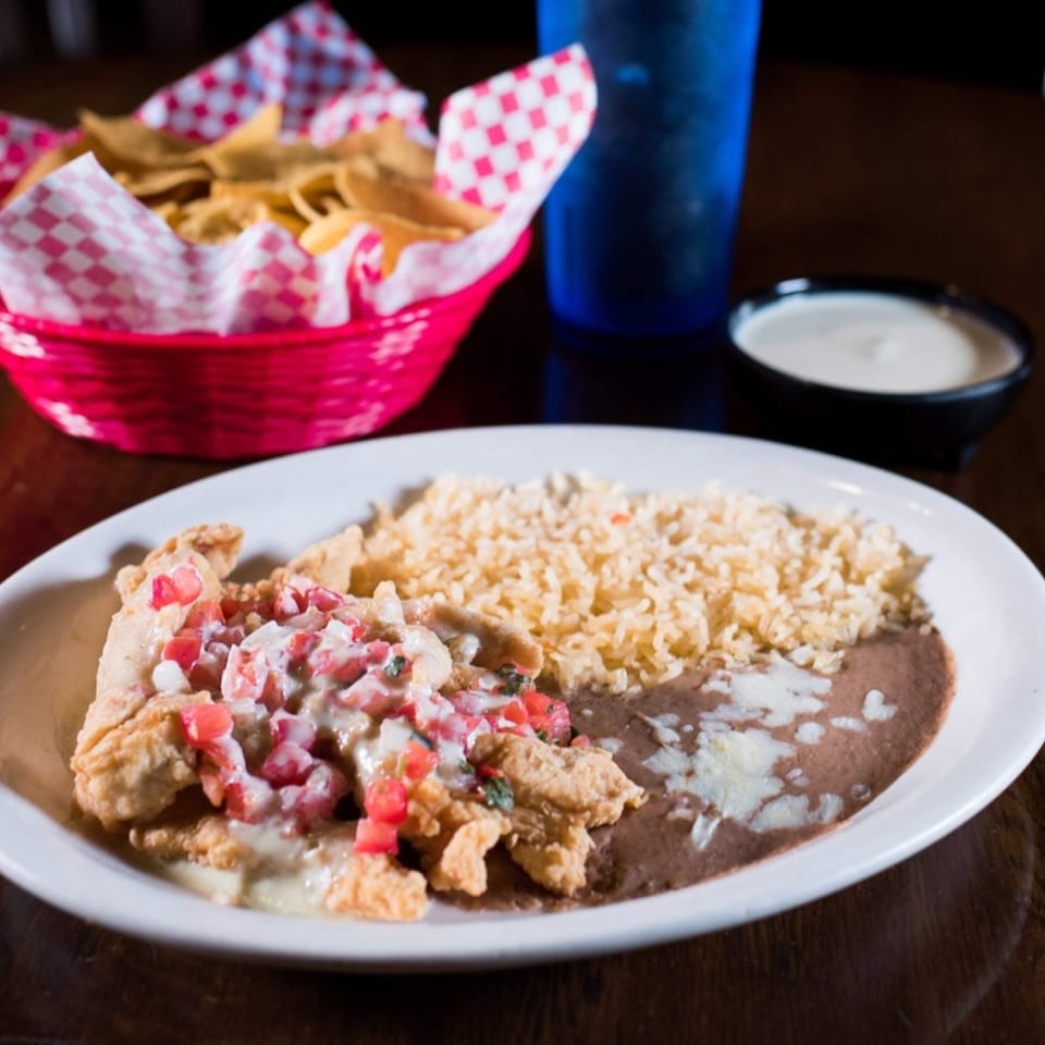Fried chicken with pico de gallo, rice, and refried beans, with chips and dip in the background.