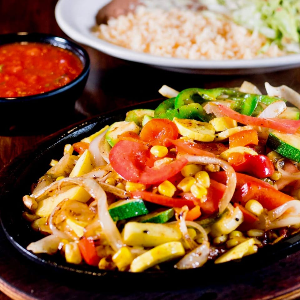 Sizzling fajita vegetables, including bell peppers, onions, zucchini, corn, and tomatoes, on a hot plate.